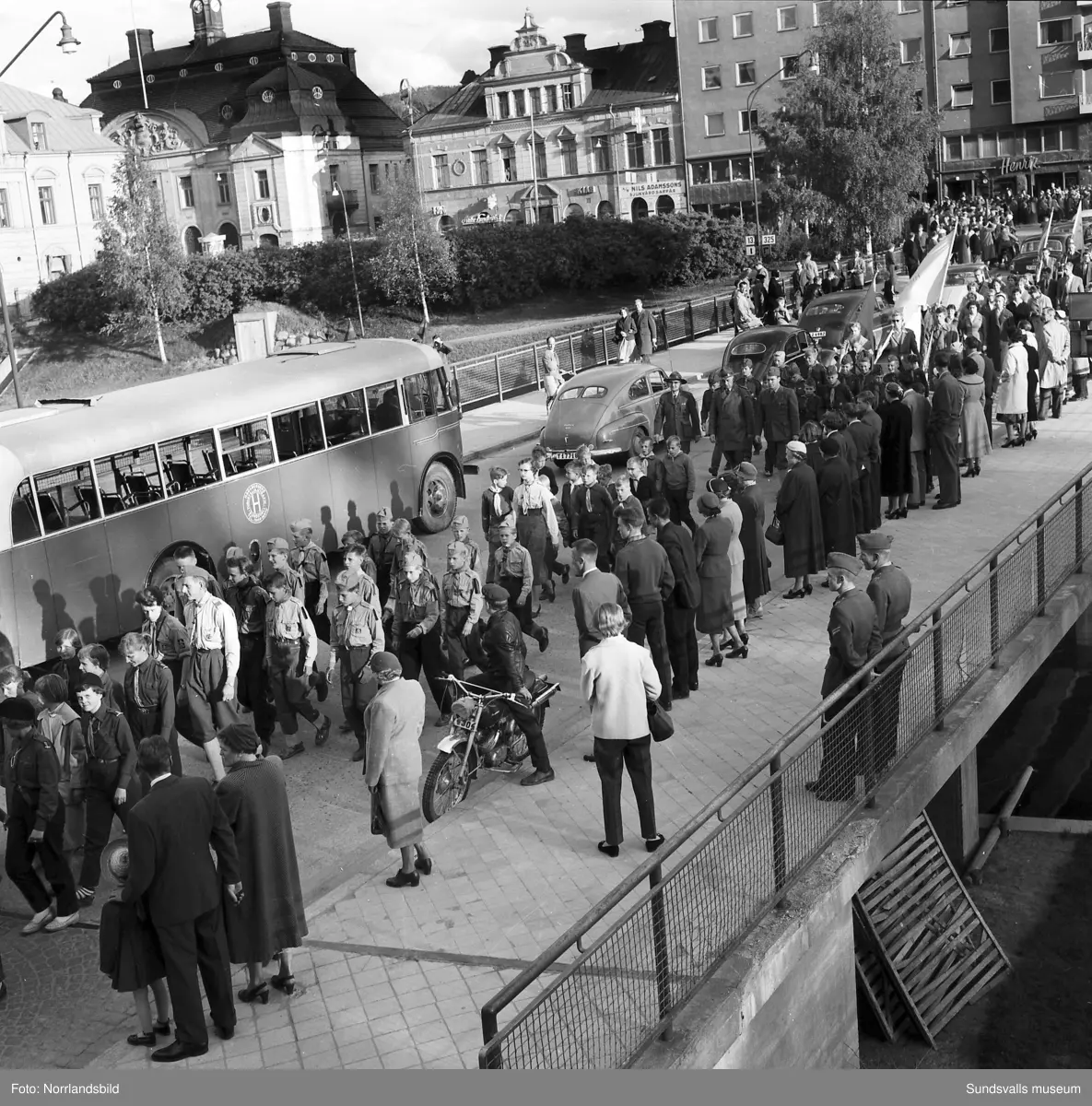 En stor serie bilder från firandet av Svenska flaggans dag 1956. Parad på Storbron med paradorkester, militär, scouter och mängder av olika föreningar och förbund. Därefter högtidlig uppställning i Idrottsparken med bland annat tal, körsång, gymnastikuppvisning och folkmusik.