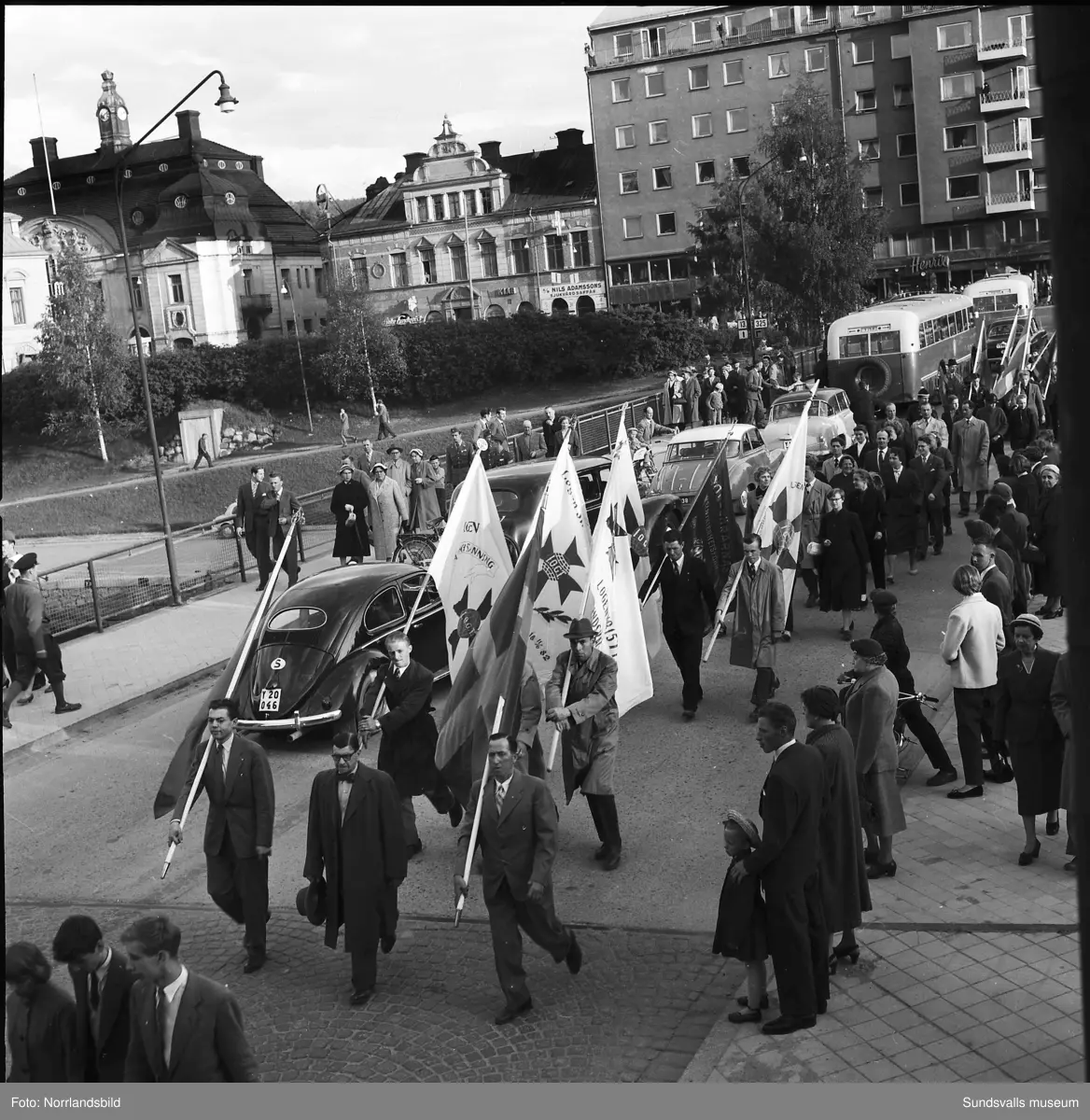 En stor serie bilder från firandet av Svenska flaggans dag 1956. Parad på Storbron med paradorkester, militär, scouter och mängder av olika föreningar och förbund. Därefter högtidlig uppställning i Idrottsparken med bland annat tal, körsång, gymnastikuppvisning och folkmusik.