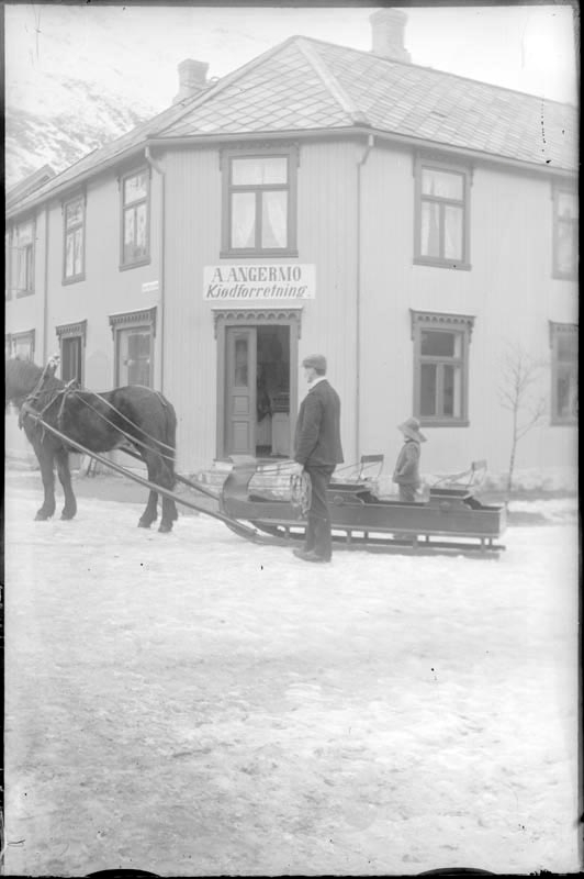 Utenfor A. Angermo kjøttforretning. Mann med hest med slede med lite barn i.