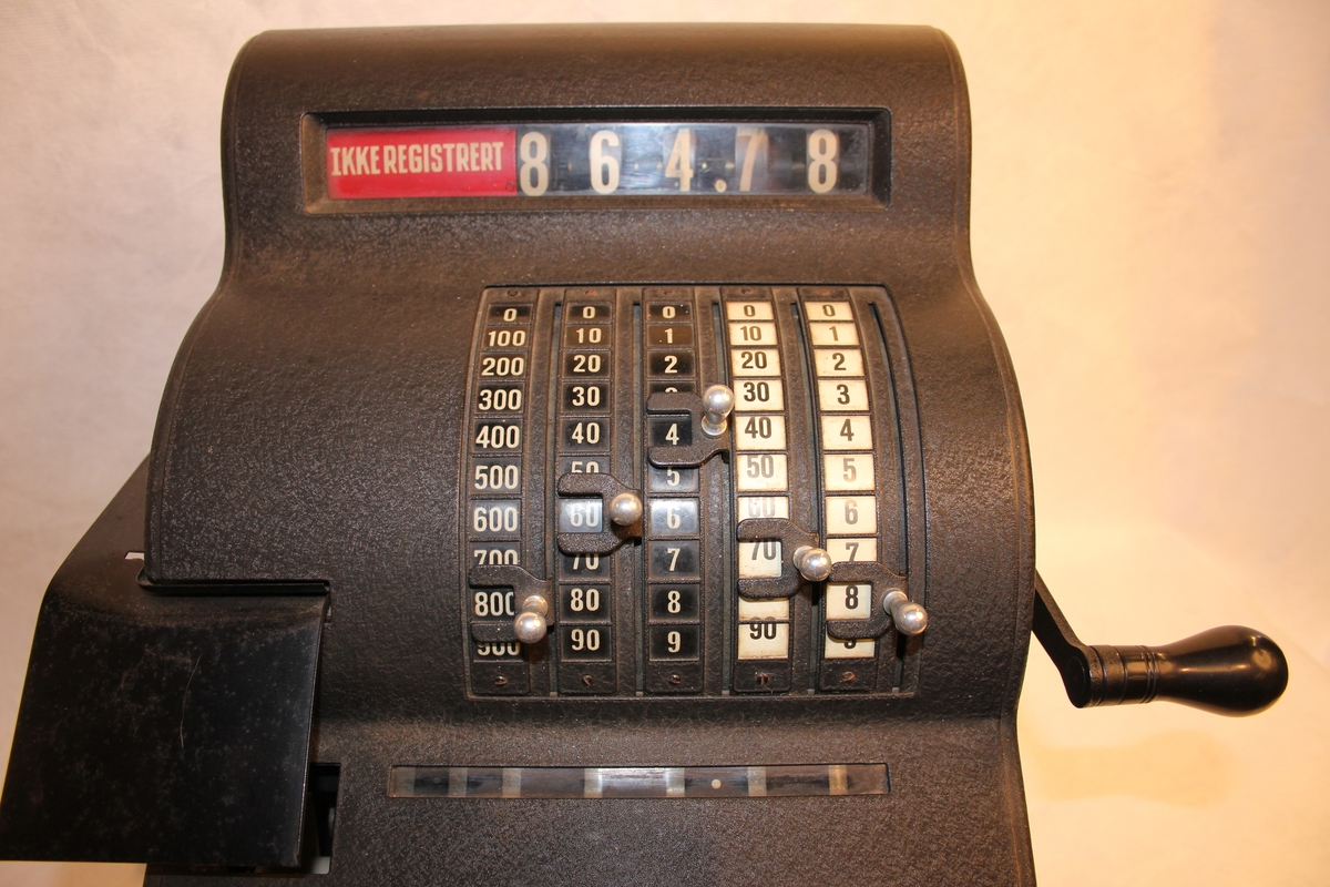 Kassa-apparat med sokkel, pengeskuff og sveiv, 5 skalaer for kr. og øre. Vindauga for kronesum framme og bak. 5 knottar. Nøkkel til skuff.