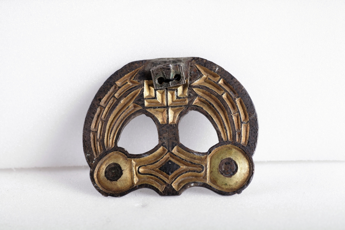 Sølvbeslag og en liten del av dens festeinnretning, forgylt med nielloinnlegg, som type R. 200 d fra eldre jernalder. Utsmykning til sverdslire. Funnet i en gravhaug på gården Mellom-Kvem.