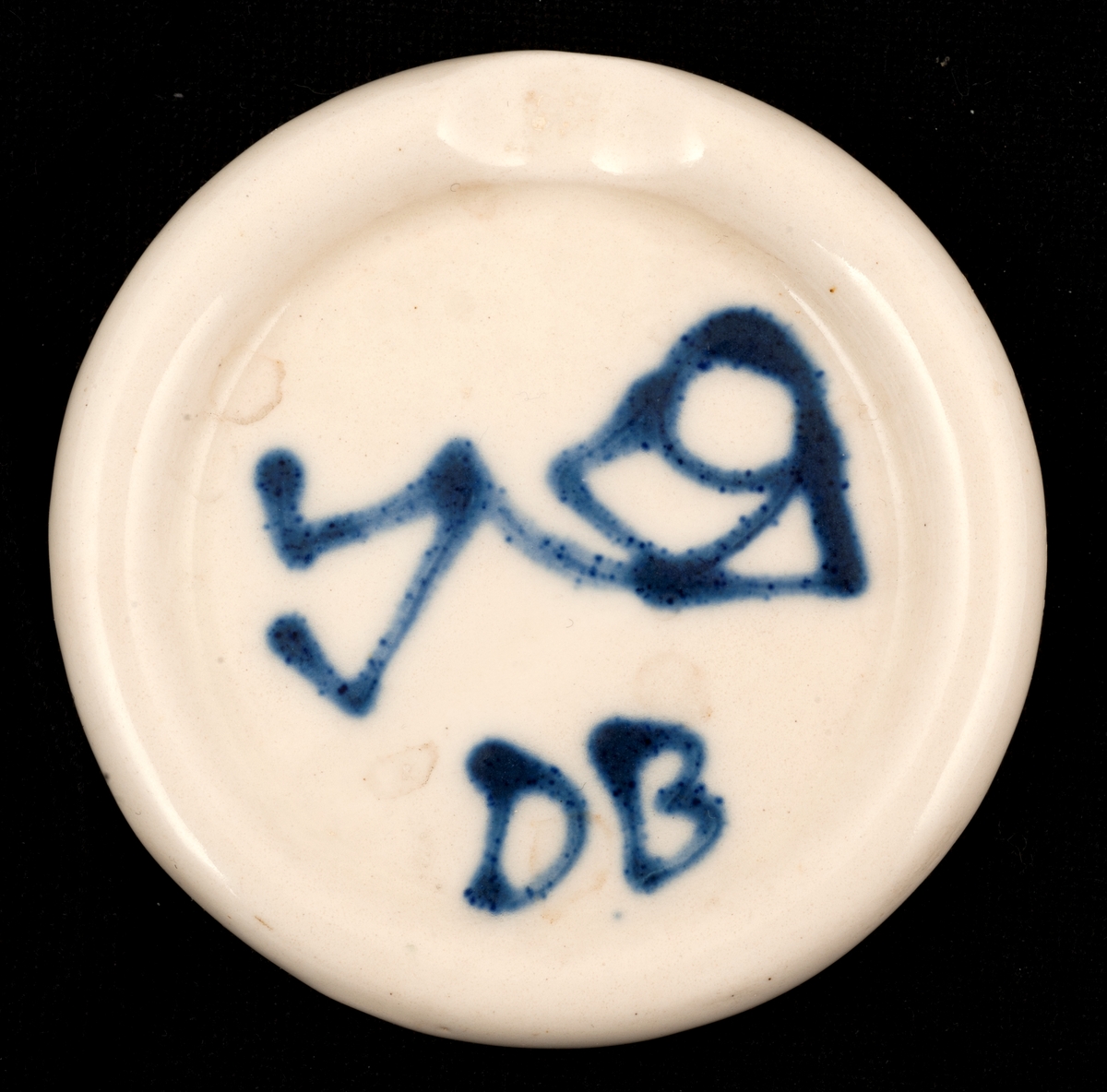 Rundt, hvitt askebeger, påmalt en liggende figur og initialene DB.