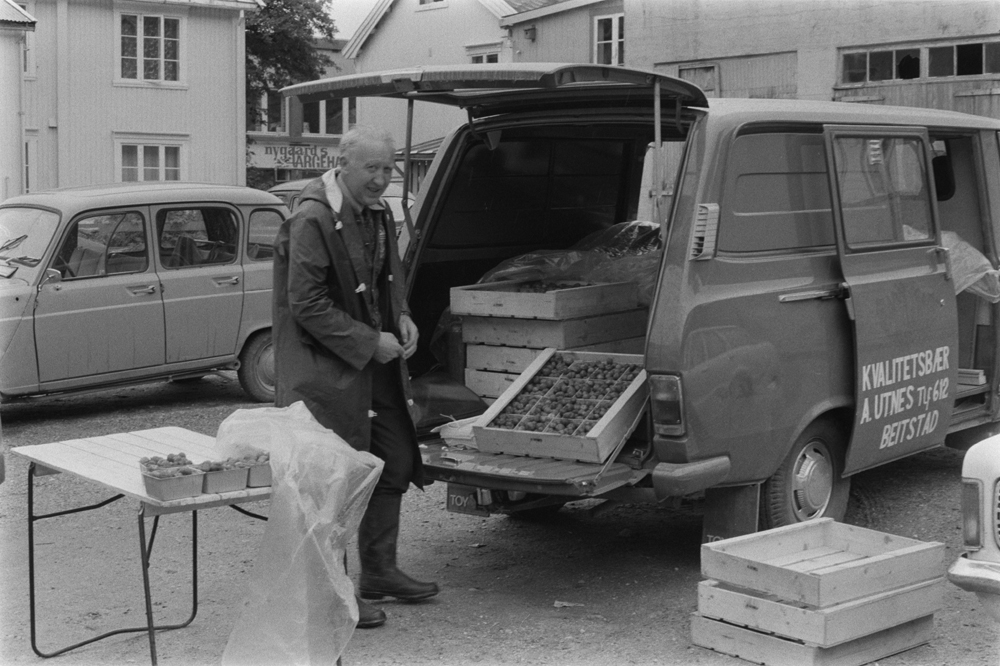 Jordbærsalg på torvet i Mosjøen. Mann som gjør klart til salg.Huset til venstre (C.M.Havigsgt. 21) er revet, huset til høyre for Nygaards Farvehandel som vi ser i C.M.Havigsgt. er Wangens Bakeri. De to andre bygningene t.h. ligger i bakgården  mellom Strandgt/C.M.Havigsgt.
Bildet er tatt fra tomta der Gammeltelegrafen sto. (Fokusbygget, Lilletorget etc.)