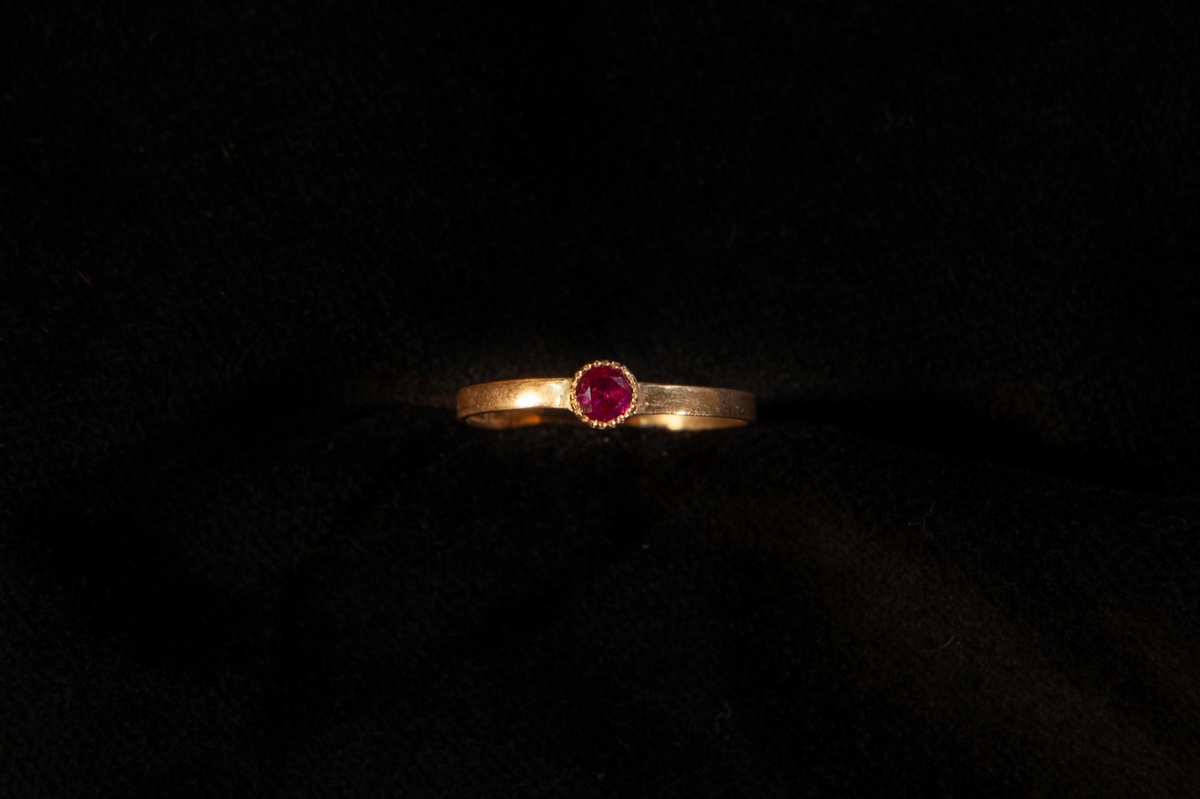 Smal guldring med infattad ljusröd sten (troligen simili (glas), möjligen rubin(?). En tillhörande rund smyckesask i papp.
