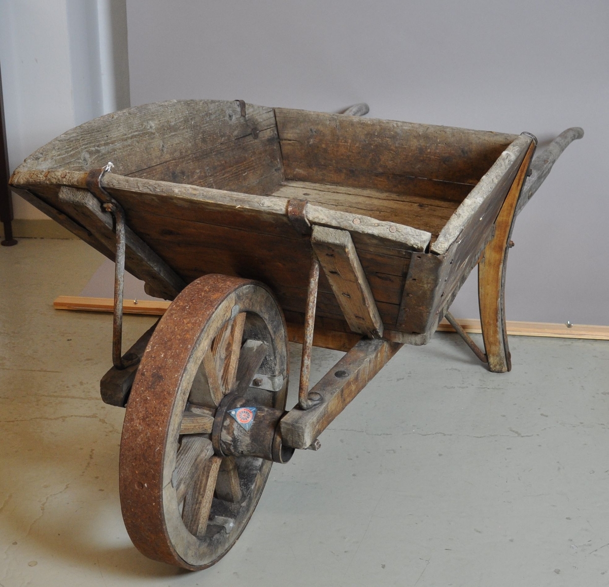 Trillebår av tre og jern. Trillebåren har en ett hjul med påfestet ring av jern, to håndtak og to støttebein. Kassen har utbøyde og avrundede sideplater. I hjørnene av kassen er det armeringer av jern.