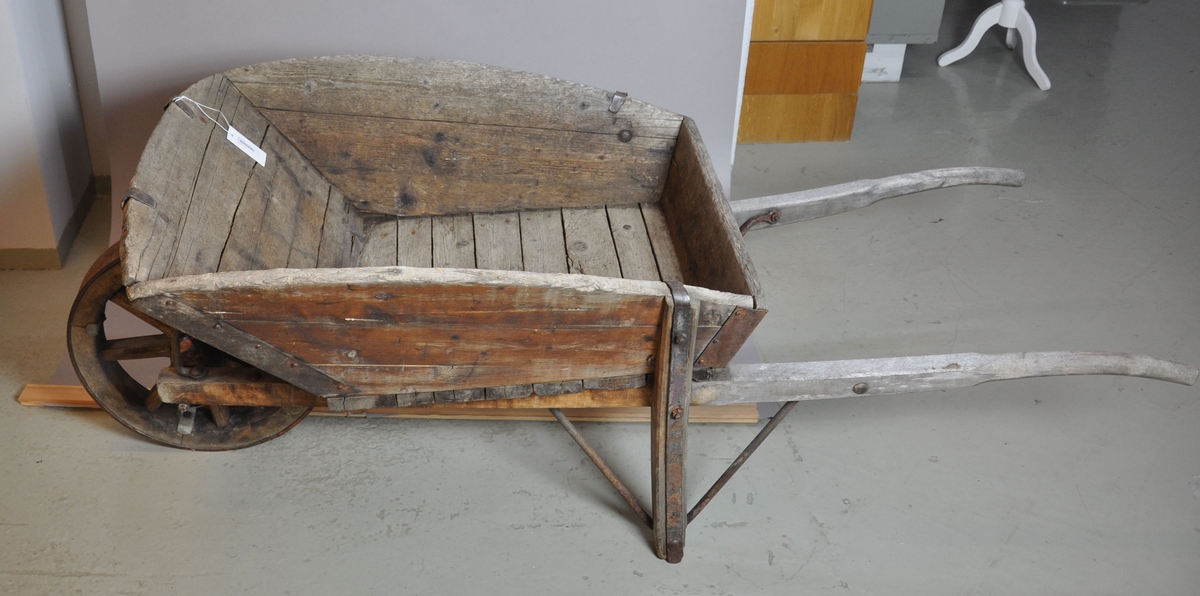 Trillebår av tre og jern. Trillebåren har en ett hjul med påfestet ring av jern, to håndtak og to støttebein. Kassen har utbøyde og avrundede sideplater. I hjørnene av kassen er det armeringer av jern.