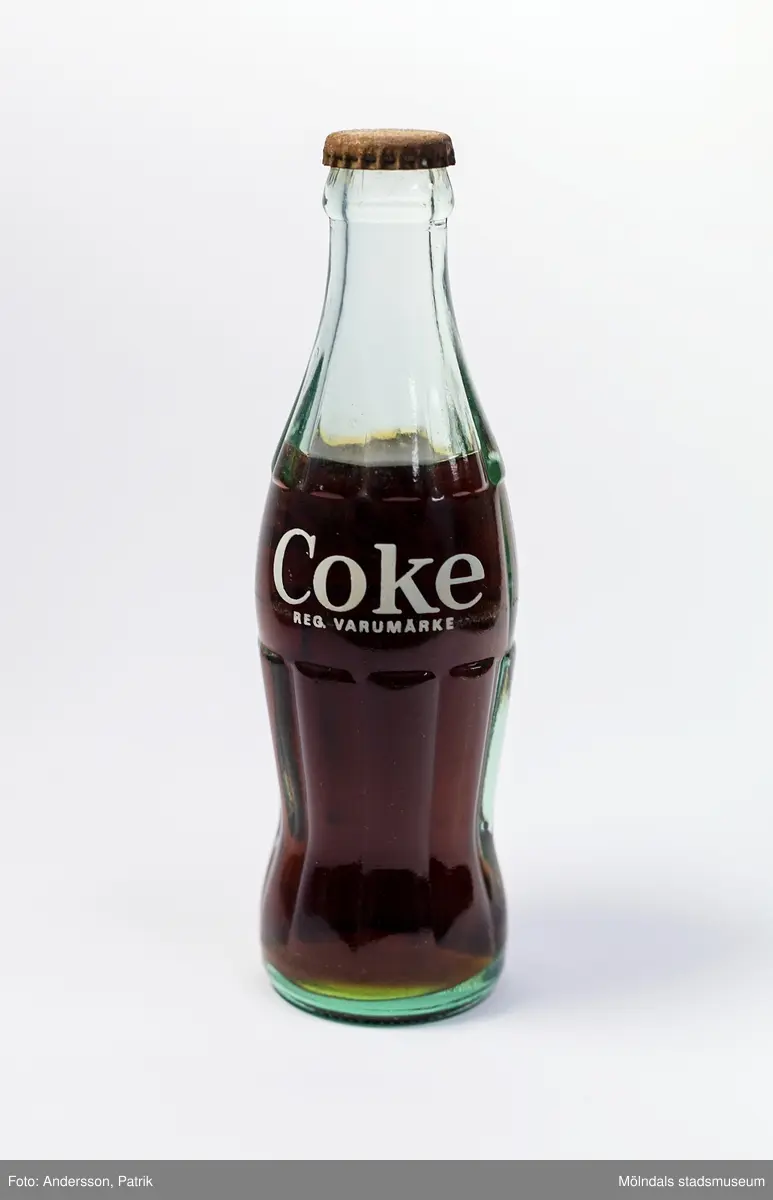 Coca-Cola i glasflaska från slutet av 1980-talet. Flaskan är tillverkad i grönaktigt glas.
Den 23 april 1985 ändrade Coca-Cola receptet på sin dryck som därmed fick namnet "New Coke". Det nya receptet blev ett totalt fiasko. Efter påtryckningar från konsumenterna återupptogs därför det gamla receptet redan i juli samma år, men då under namnet "Coca-Cola Classic". När "Coca-Cola Classic" kom tillbaka, sålde den redan från början mer än "New Coke". 

Glasflaskans form är inspirerad av kakaobönan. Det geniala med flaskan är dess greppvänliga form med dess särpräglade utseende som gör att man känner igen den även om den greppas i totalt mörker. Glaset splittrades heller inte om flaskan tappades i marken. I början tillverkades flaskorna i färgerna blått, grönt och glasklart glas.

Glasingenjören Alexander Samuelson som formgav flaskan har koppling till Sverige. Han föddes nämligen i Kungälv den 4 januari 1862. Under sina ungdomsår arbetade han på Surte glasbruk. Vid 21 års ålder flyttade han med sin storebror till USA, där han fortsatte att arbeta på olika glasbruk och blev så småingom chef. I början av 1910-talet anordnades en tävling för att designa en flaska till Coca-Colan. Tävlingen vanns av Alexander och hans glasbruk. 1915 tog Alexander Samuelson patent på denna geniala utformning av Coca-Colaflaskan, en av världens mest välkända varuförpackningar.

Eftersom livsmedelslagen i Sverige hade förbud mot koffein och fosforsyra, kunde inte Coca-Colan börja säljas i Sverige förrän 1953 då förbudet upphäves. Det var då Pripps bryggerier som tog hem lincensen för att börja tillverkningen av den amerikanska drycken i Göteborg, Malmö och Sundbyberg. Coca-Colan hade först svårt att konkurrera med storheter som Sockerdricka, Champis, Festis och Zingo. Det var först i slutet av 1970-talet som försäljningen ökade ordentligt.

Enligt inkommen kommentar: Enligt inskriften är flaskan tillverkad 1968 på Hammars glasbruk. Men drycken kan ju vara från 1980-talet.