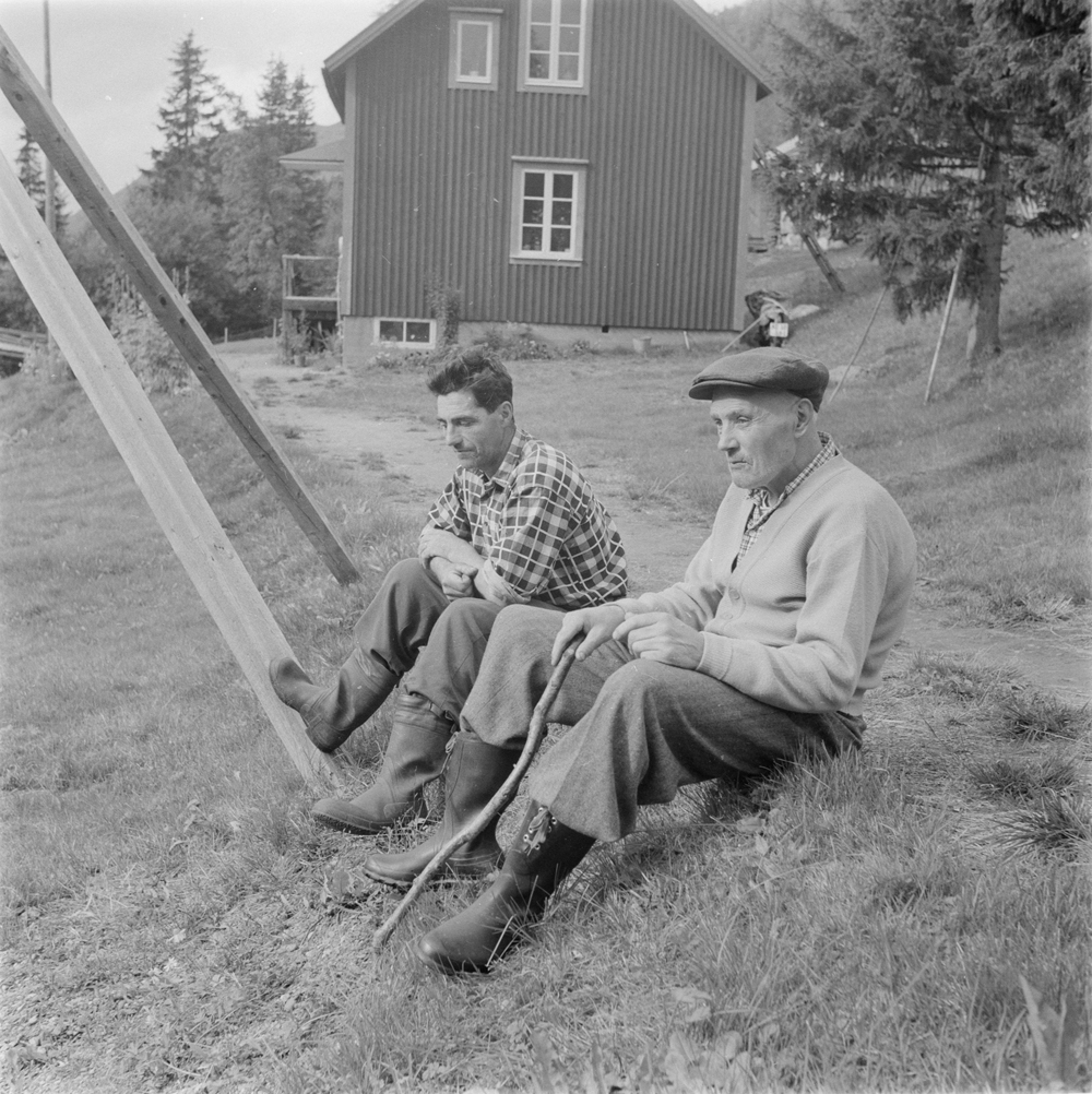 To menn sittende i graset. Hus i bakgrunnen.
Fra Skarmodalen?