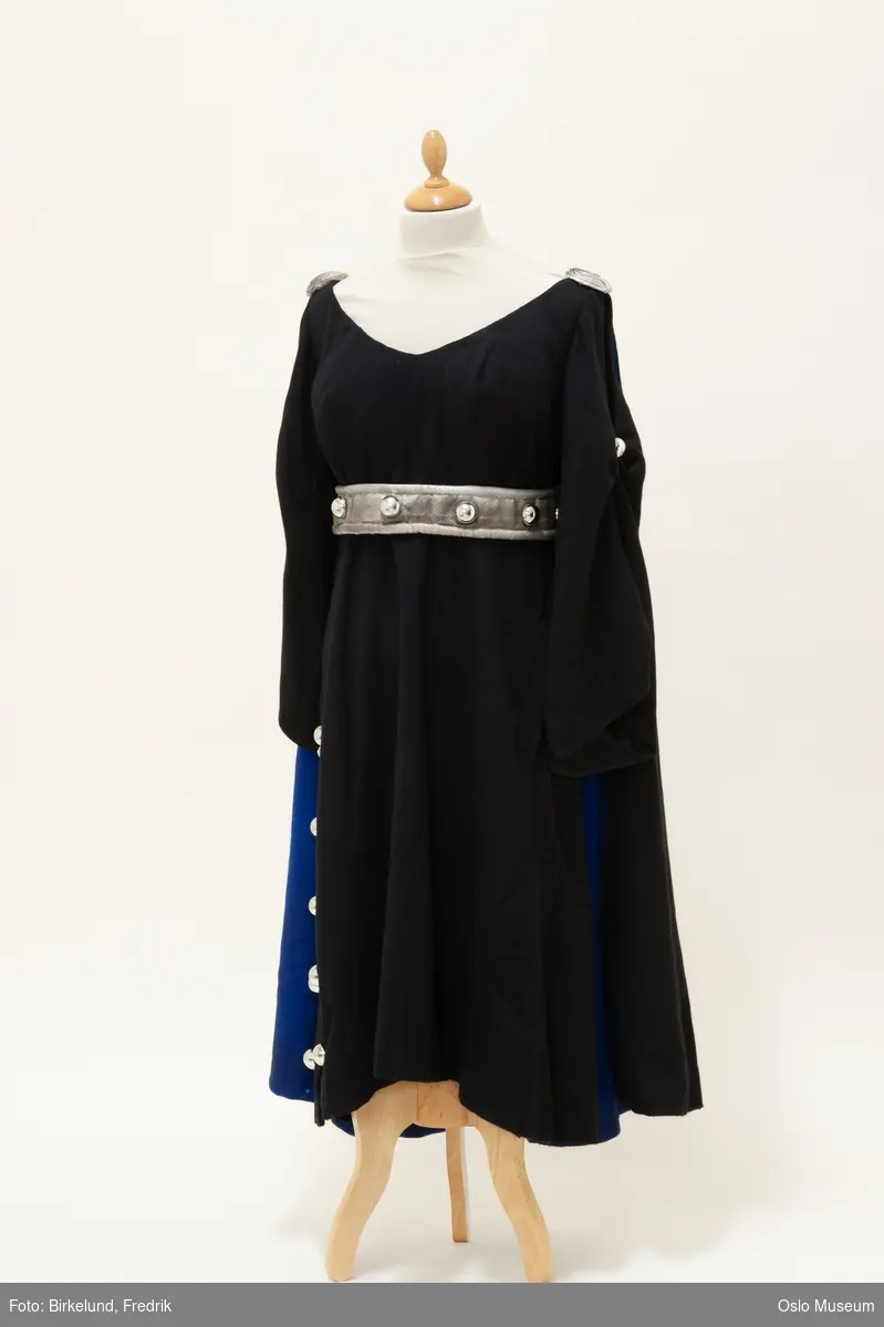 A: Lang, svart og blå kappe i ull med påsydd dekor
B: Sølvfarget tekstilbelte med påsydd dekor
C: Søvlfarget tekstilbelte med påsydd dekor
