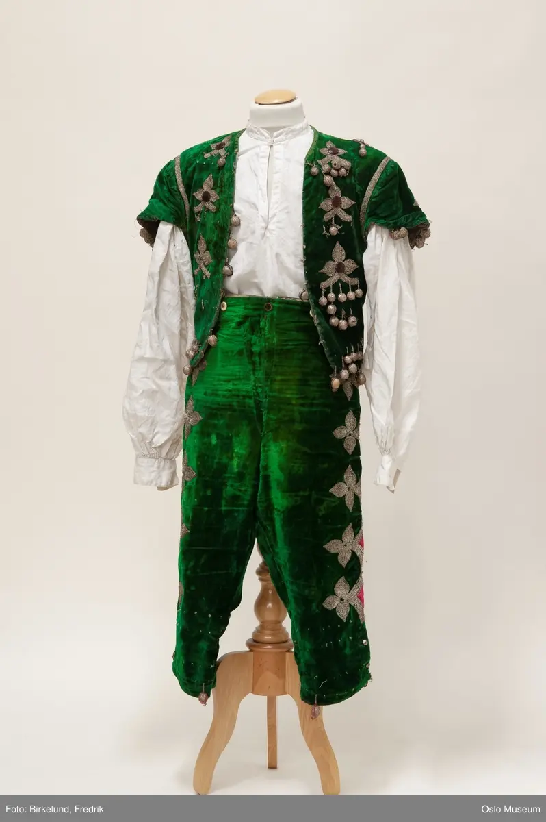 A: vest, grønn bunn med broderte blomster i metallfarget tråd. Pyntet med rød plaststeiner. 
B: bukse, grønn bunn med blomster brodert i metallfarget tråd langs kanten. Rosa dekor. 
C: skjorte, hvit lin. 
D: løse deler
E: vest