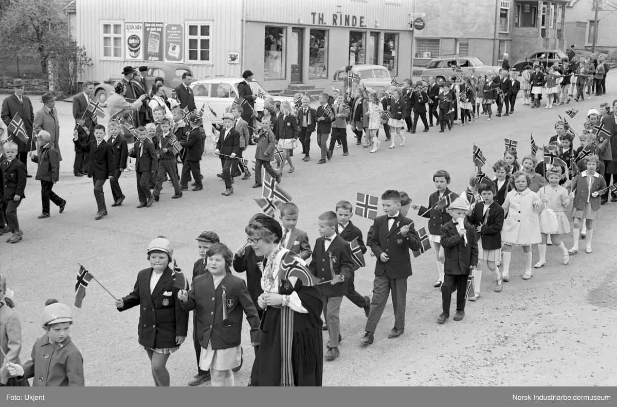 Barnetog nedover gate. På kolonialforretningen over gaten står det "TH.RINDE". Dette er kolnialforretningen startet av Thorstein Rinde i 1929. Butikken brant ned til grunnen 24.12.2005.