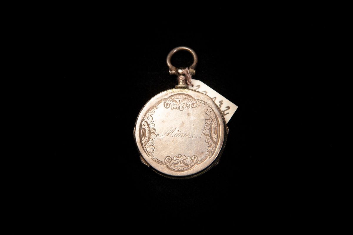 En rund medaljong av silver som går att öppna på både fram och baksida. Framtill graverat: "Minne", omgivet av snirklig bård. Baktill graverat en palmkvist lutad över ett kors. Stämplad invändigt.