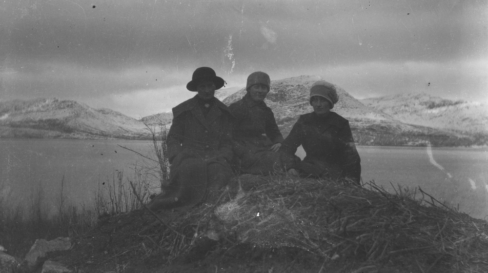 Tre kvinner sitter i gresset. Fjell og sjø i bakgrunnen. Hattfjelldal.
Oleanna Linvik, ukjent og Emelia Linvik.