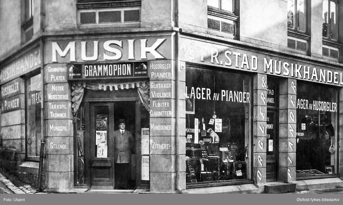 Eksteriør av A/S Fredrikstad Musikhandel i Bakgaten 6 med sin innehaver i døråpningen, ukjent person, kan det være Mathiesen? 
Navn på utstillingsvindu: Mathiesen. Ukjent tidspunkt, ant. ca. 1910. 

Reklame for følgende musikkinstrumenter: piano, husorgel, fiolin, fløyte, klarinett, mandolin, gitar, trekkspill, munnspill, zither, trommer, samt grammofon.

I følge "Adressebok for Østfold fylke : med skatteligninger. 1937 Vol. 3" har Fredrikstad Musikkhandel adresse Bakgaten 6, så nr. på veggen stemmer.