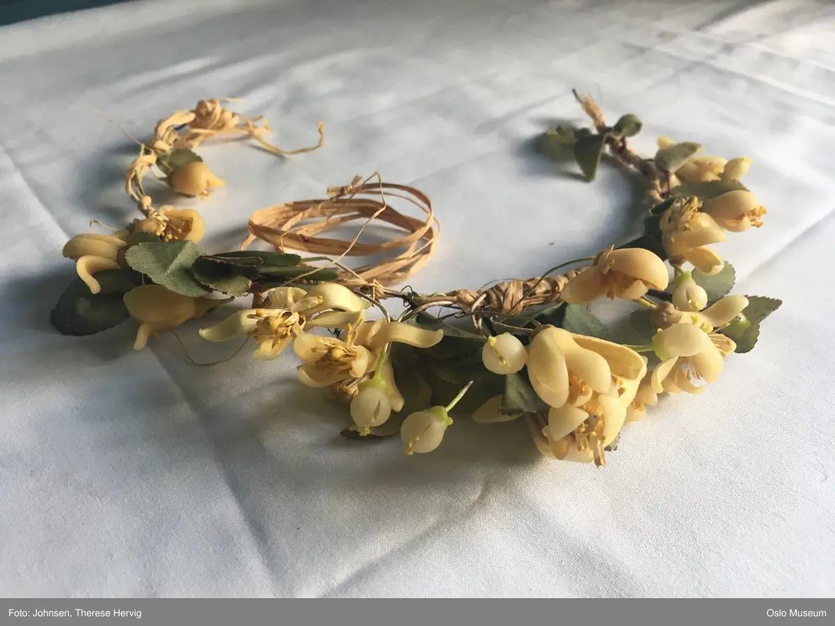 A: Rett fasong fra byste ned over hofte og lårparti. Løsere i skjørtet nedenfor hofte/lår.  Ermeløs med trykknapper i siden. Øvre tøylag er gulhvit blonde med blomsterornamentikk.  Blonden er festet og henger over en rosa underkjole. Det rosa stoffet skinner gjennom og gjør kjolen blek rosa.
B: hårkrans