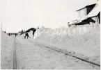 Vy på spår och stationshus med snövallar och pågående snöröjning.