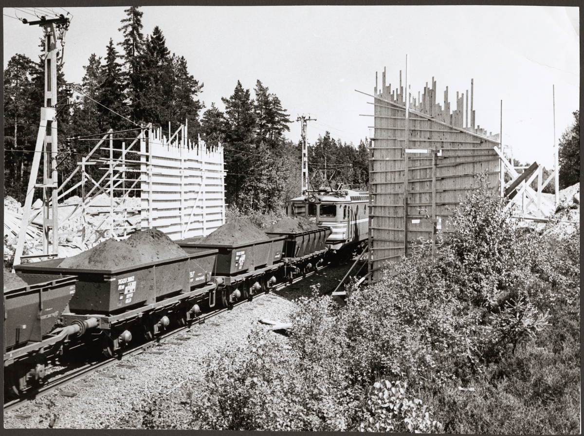 Malmtåg med bland annat Trafikaktiebolaget Grängesberg - Oxelösunds Järnvägar, TGOJ M30 3062 draget av ett ellok under påbörjat bygge av viadukt. Bygget sker vid Snarabacken för den nya Kvicksundsleden, väster om Snara banvaktstuga.