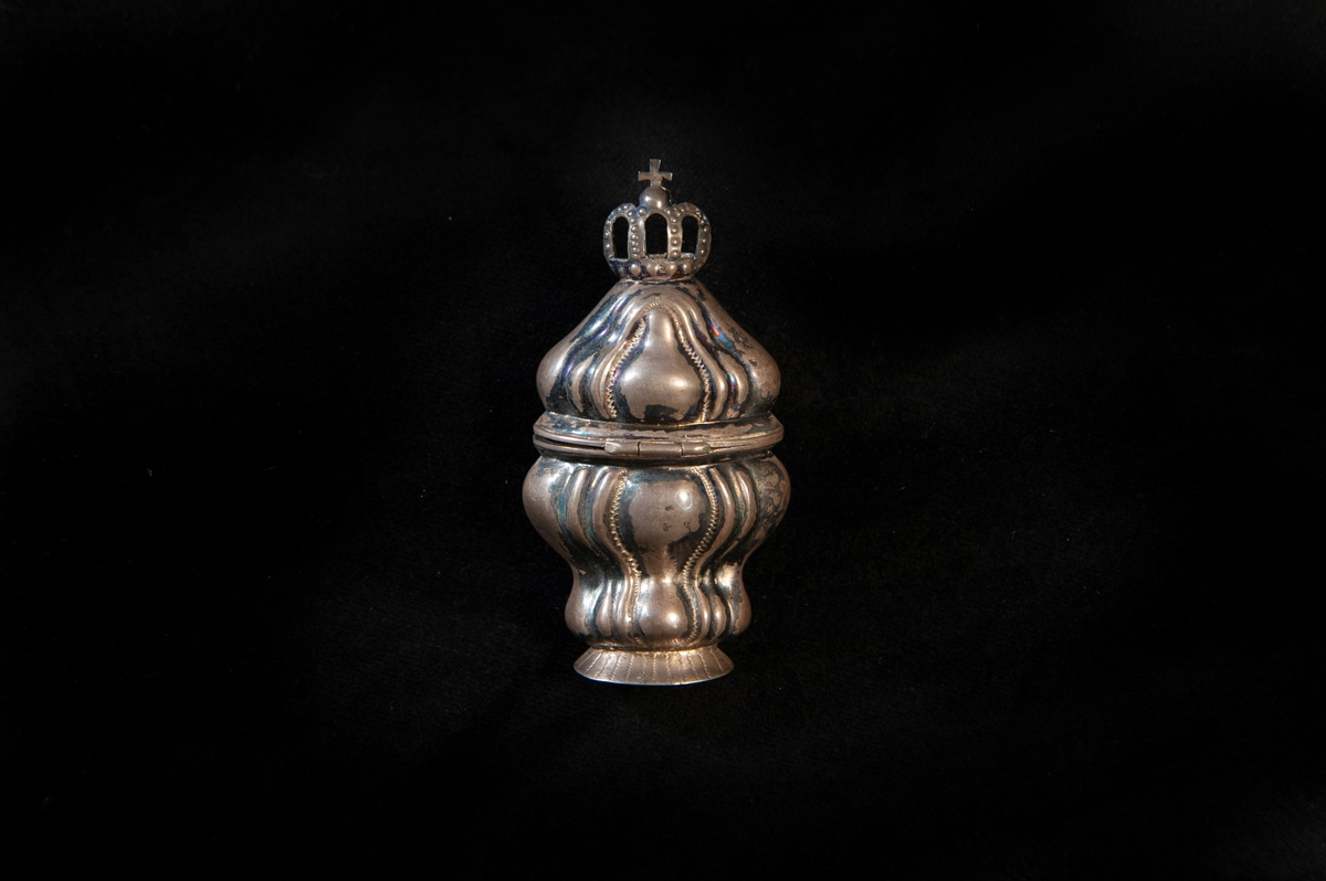 Hög dosa av silver i rokoko. Dekorerad med så kallade "knäckta åsar" i relief, och högst upp på locket en genombruten krona med kors. Utländska stämplar i botten. Troligen dansk tillverkning. Jämför med JM 18 742 och 19 952.
