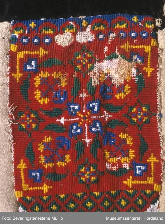 Rektangulært broderi på kvadratisk bakstykke. Plattsaum (halve kross-sting) i fleire retningar sydd på stramei. Nokre usydde og utslitte område. Raud hovudfarge. Diverse geometriske figurar og ei slags åttebladrose i midten. Bord nede  og oppe. Broderistoffet går litt nedom den nederste borda. Svart kant oppe. Alt er sydd på eit ullstoff vove i kypert. Fleire hol.