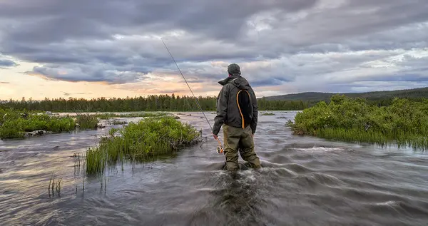 Mann med fluefiskestang og vadre som står i en elv