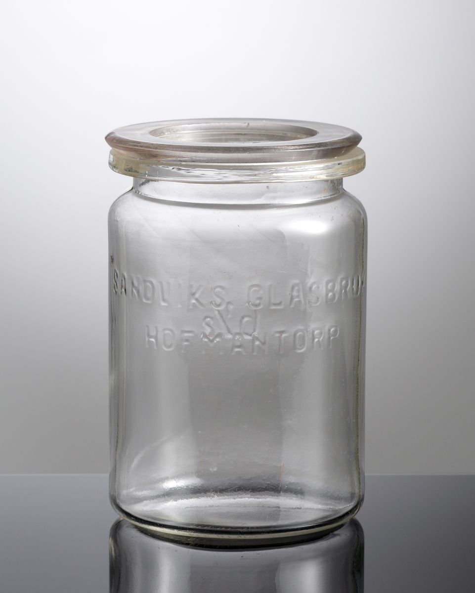 Konserveringsburk med lock.
Tillverkad på Sandviks glasbruk.
Cylindrisk, kraftig krage, planslipad matt mynning.
Upphöjd relief med text: "Sandviks glasbruk Hofmantorp"
Blåst i tvådelad form. Locket pressat.