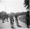 Sex män på cykel i riktning mot fotografen. På Visingsö med kungsladugården till vänster i bakgrunden