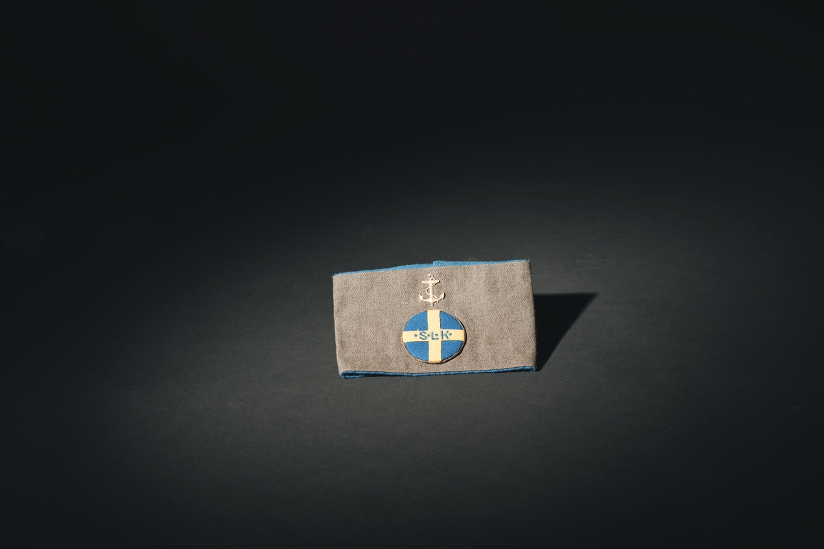 Armbindel för marinlotta. Rektangulärt tygstycke, dubbelt. Framstycket gråbrungrönt, bakstycket mellanblått. Bakstycket bildar blå passpoal runt det gråbrungröna stycket. I mitten på framstycket cirkelrunt emblem i tyg: Riksförbundet Sveriges lottakår: blå botten, gult kors med broderade bokstäver, blå: " . S . L . K .". Lagd gul metallpåsytt. Ovan emblemet gul metallankare fastsytt: flottans emblem.

:1 ihopsydd i kortändarna.