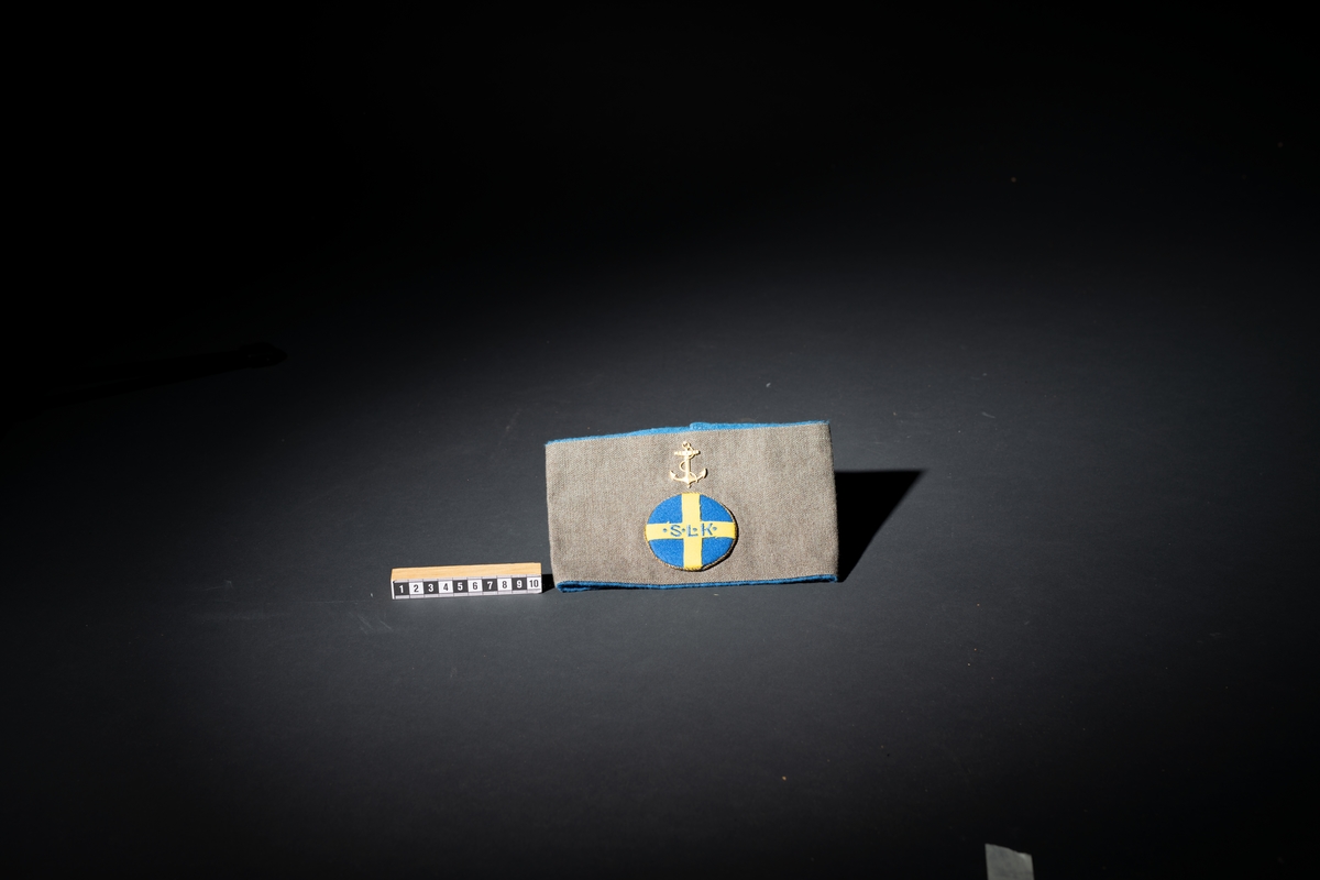 Armbindel för marinlotta. Rektangulärt tygstycke, dubbelt. Framstycket gråbrungrönt, bakstycket mellanblått. Bakstycket bildar blå passpoal runt det gråbrungröna stycket. I mitten på framstycket cirkelrunt emblem i tyg: Riksförbundet Sveriges lottakår: blå botten, gult kors med broderade bokstäver, blå: " . S . L . K .". Lagd gul metallpåsytt. Ovan emblemet gul metallankare fastsytt: flottans emblem.

:1 ihopsydd i kortändarna.