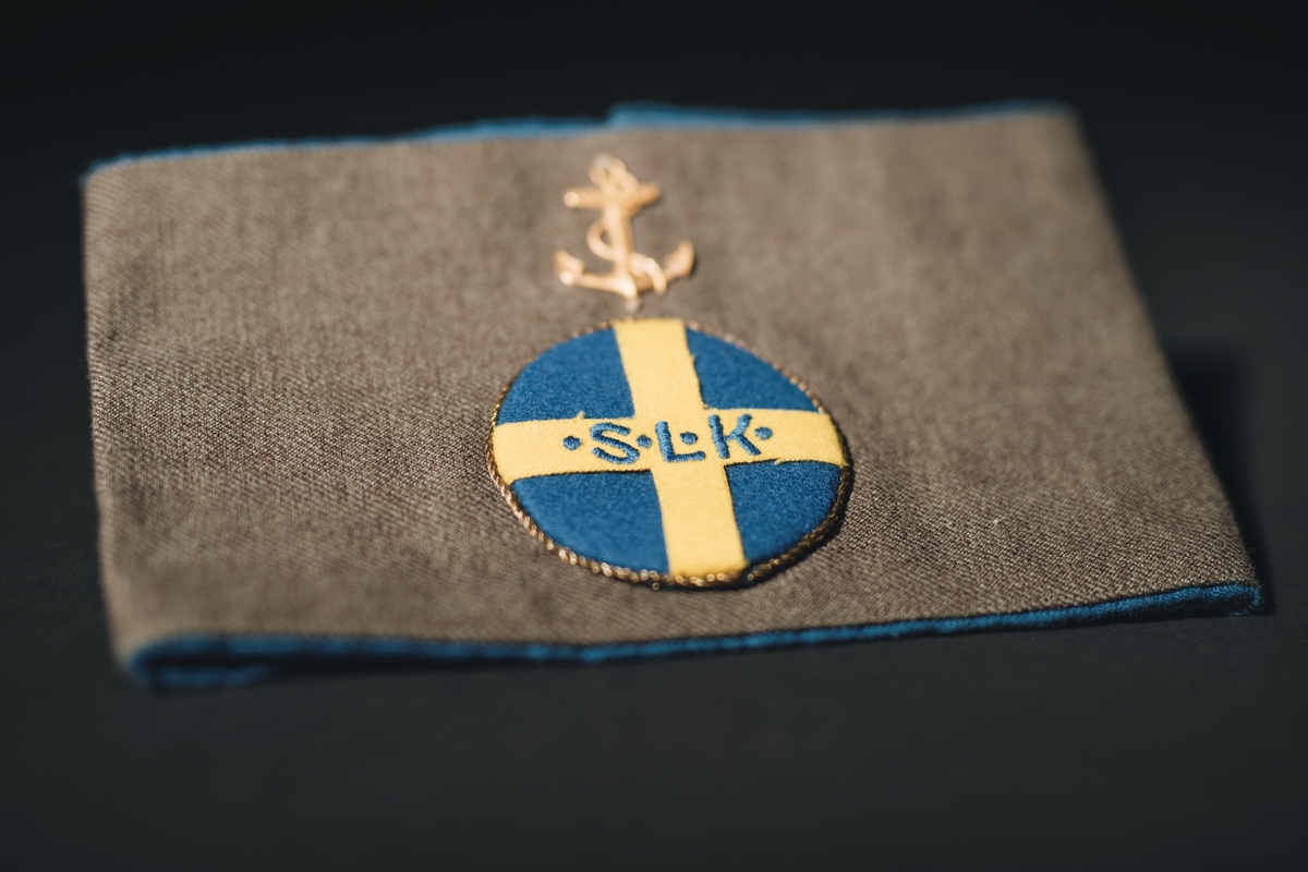 Armbindel för marinlotta. Rektangulärt tygstycke, dubbelt. Framstycket gråbrungrönt, bakstycket mellanblått. Bakstycket bildar blå passpoal runt det gråbrungröna stycket. I mitten på framstycket cirkelrunt emblem i tyg: Riksförbundet Sveriges lottakår: blå botten, gult kors med broderade bokstäver, blå: " . S . L . K .". Lagd gul metallpåsytt. Ovan emblemet gul metallankare fastsytt: flottans emblem.

:1 ihopsydd i kortändarna.