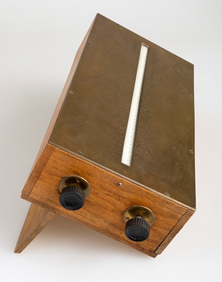 Kubikkberegningsapparat, levert av produsenten F. A. Krog i Skovveien i Kristiania, antakelig tidlig på 1900-tallet.  Apparatet er formet som et rektangulært skrin, utført i eiketre, men med "lokk" av ei messingplate.  Langs midtaksen på dette lokket er det ei lang, smal, rektanuglær spalte med glass under.  Gjennom denne ser vi ned på en tabell.  Tabellen er trykket på en papirrull, som er forankret på to valser nede i skrinet.  Tabellen kan beveges ved hjelp av dreiehjul på skrinets endeflater, to på hver side, ett i hver ende av de nevnte valsene.  Lengst til høyre i tabellvinduet finner vi "klassifiseringsinformasjon" - angivelser av stokklengder, diametere og salgsklasser.  Langs den øvrige delen av vinduet kan man lese av kubikkmeterangivelser for fra 1 til 70 stokker med samme dimensjoner og klassespesifikasjon.  Avlesinga skjer fra en tolinjet tabell på den nevnte papirrullen inne i skrinet, og med stokketallesangivelser på lokket - 1-35 stokker på oversida av vinduet og 36-70 stokker på undersida.  Det nevnte emaljerte lokket, som er hvitt med svarte tall og bokstaver, er skrudd fast til toppen av skrinet.  Det er grunn til å tro at valsene med tabellen må være montert før dette ble påsatt.  Skrinet er ellers lagd av høvlet eiketre, og den holdes sammen av de nevnte skruene i lokket og spiker fra botnstykket.  På standflata er det også hengslet en klaff, som kan vippes opp, slik at lokket skrår mot brukeren når apparatet brukes på et bord eller et annet plant underlag. 