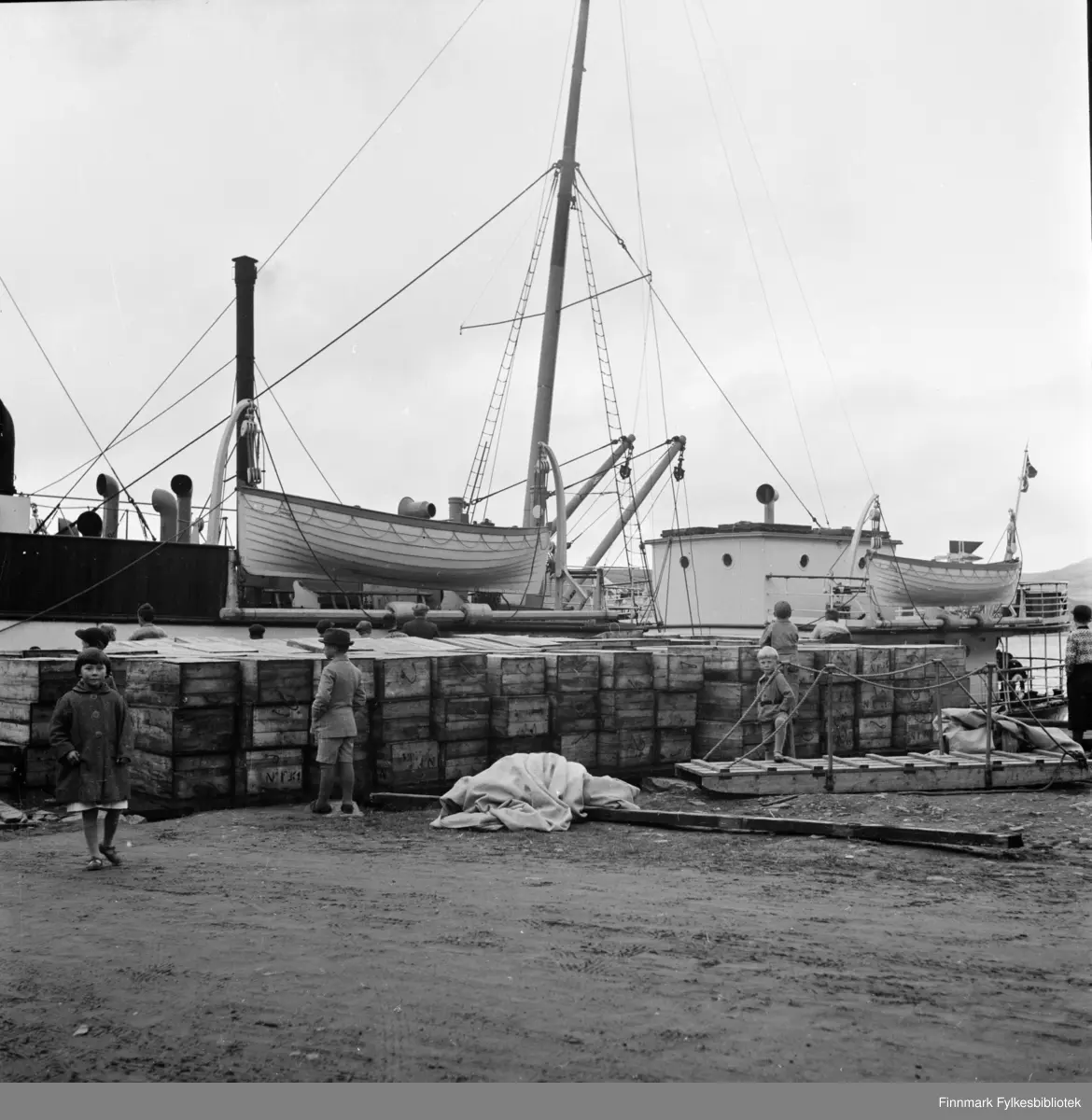 Kjøllefjord i 1940. Ved kaia ligger Bergenskes hurtigruteskip D/S IRMA. En av kaiene i Kjøllefjord med oppstablede kasser, sannsynligvis med saltfisk. Kassene er merket NF. NF sto for Nils Foldal. I forgrunnen ses flere barn, blant annet Jon Svendsen som står med ett bein på en landgang til høyre i bildet. I bakgrunnen ligger en båt til kai. 

Nils Foldal kom fra Follestaddalen ved Ørsta. Han kom til Kjøllefjord i 1912 og fikk arbeide hos brødrene Aarsether som den gang disponerte Horst & Christensenbruket. I 1914 kjøpte han en eiendom av Sveen der det var noe bebyggelse. Han startet med kjøp av fisk, bl.a. sild samt tranproduksjon, restaurerte bygninger og forlenget kaia, og i 1927 overtok han som dampskipsekspeditør for Vesteraalens Dampskibsselskab som hadde tre hurtigruter og flere godsbåter i Kjøllefjord. Foldal-familien flyttet fra Kjøllefjord til Ålesund i 1932. 

I bakgrunnen sees Pedersen og Nissens fiskebruk til venstre og til høyre ligger Bjørgvik hotell. Søstrene Bjørgvik drev hotellet i Kjøllefjord.

Utdrag fra tekst hvor Foldalbruket forteller om Nils Foldals virksomhet: 

«Foldalbruket ble bygget opp igjen etter krigen. Frem til 1959 hadde Foldalbruket også eksepdisjonskai med hurtigruteanløp. Foldalbruket var tidligere et fiskebruk med tradisjoner tilbake til 1900-tallet. Sunnmøringen Nils Andreas Foldal ankom Kjøllefjord i 1917 og fikk jobb ved Aarsæthers trandamperi. Her lærte han tranproduksjon og tørrfiskproduksjon og etablerte så fiskebruket Foldalbruket i 1917 og de drev helårsdrift. Det ble kjøpt fisk til både salting og tørking. Kassene med saltfisk på bildet er sannsynligvis iferd med å bli lastet om bord. Den største fisken ble flekket og saltet før den ble sendt til Sunnmørskysten og tørket som klippfisk. Resten ble sperret og tørket. Denne tørrfisken ble eksportert til både Italia og Afrika.
Nils Foldal ble ekspeditør for Vesteraalens Dampskibsselskap i 1926, og bygget i denne sammenheng ut kaia ved bruket slik at båtene kunne legge til. Tidlig på høsten 1927 gikk den første hurtigruta til kai i Kjøllefjord. Foldal ble etterhvert også ekspeditør for Ofoten og for Nordenlandske Dampskibsselskap. Han var en populær mann i ledelsen i disse selskapene fordi han, ifølge nevø Lars Foldal var så god å skaffe last.
– Ved en anledning sendte han 400 kasser sei med hurtigruta. Da lå skipet tungt i vannet på tur fra kai, forteller Foldal.
I de 19 årene Nils Foldal drev eget firma i Kjøllefjord fikk han utrettet mye, også utenfor bygda. Lofoten var et av stedene han drev stort. Både Svolvær, Henningsvær og Balstad var blant de stedene Foldal drev sin virksomhet i. Han eksporterte også mengder av sine produkter både til Newcastle og Frankrike i tillegg til Italia og Afrika.
Nils Foldal flyttet til Ålesund i 1933. Han hadde fått jobb som ekspeditør for Vesteraalske dampskibsselskap i Ålesund. Hans nevø Lars Rebbestad overtok som bestyrer i Kjøllefjord. Han var en av de største fiskeoppkjøpere i Kjøllefjord frem mot andre verdenskrig. Det var stor aktivitet på Foldalbruket frem mot krigsutbruddet. Hurtigrutetrafikken stanset høsten 1941, men erstatningsbåter gikk for de forskjellige selskapene ennå en tid. 

Høsten 1944, 4.-6. november brente og sprengte tyske marinesoldater hus og kaier i Kjøllefjord.»