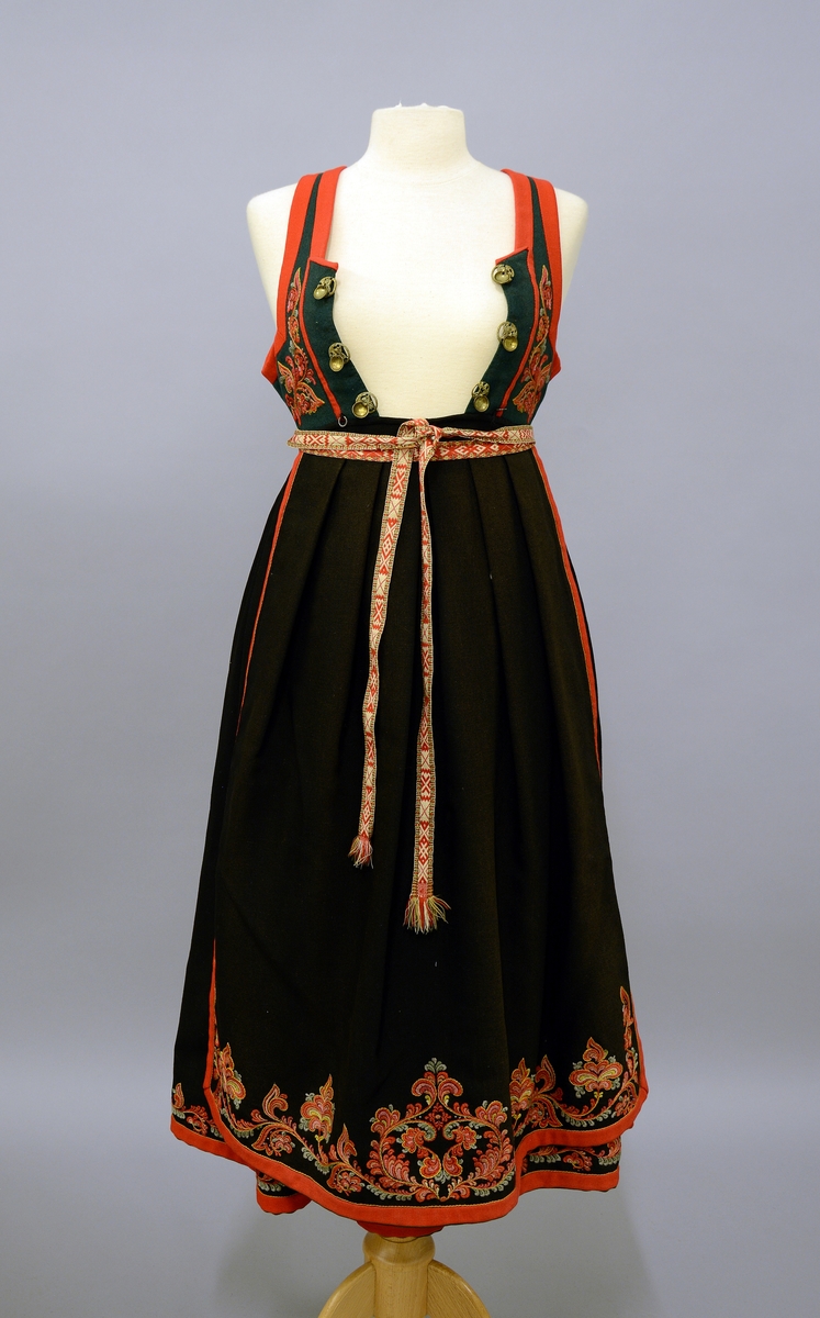 Modellbunad fra Vest-Telemark.TGM-BM.1999:0143.A-D.
B. Forkle. Sort forkle i ull med brodert bord i rosesøm nederst. Forkleet er kantet med et rødt og et grønt ullbånd. Påmontert forklebånd i grindvevteknikk i fargene rødt, hvitt, beige og grønt.