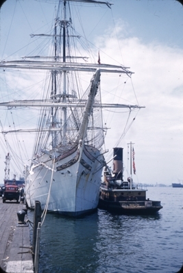 Skoleskipet STATSRAAD LEHMKUHL til kai, slepebåt fortøyd på yttersiden av fartøyet. På bryggen er en rød bil parkert.