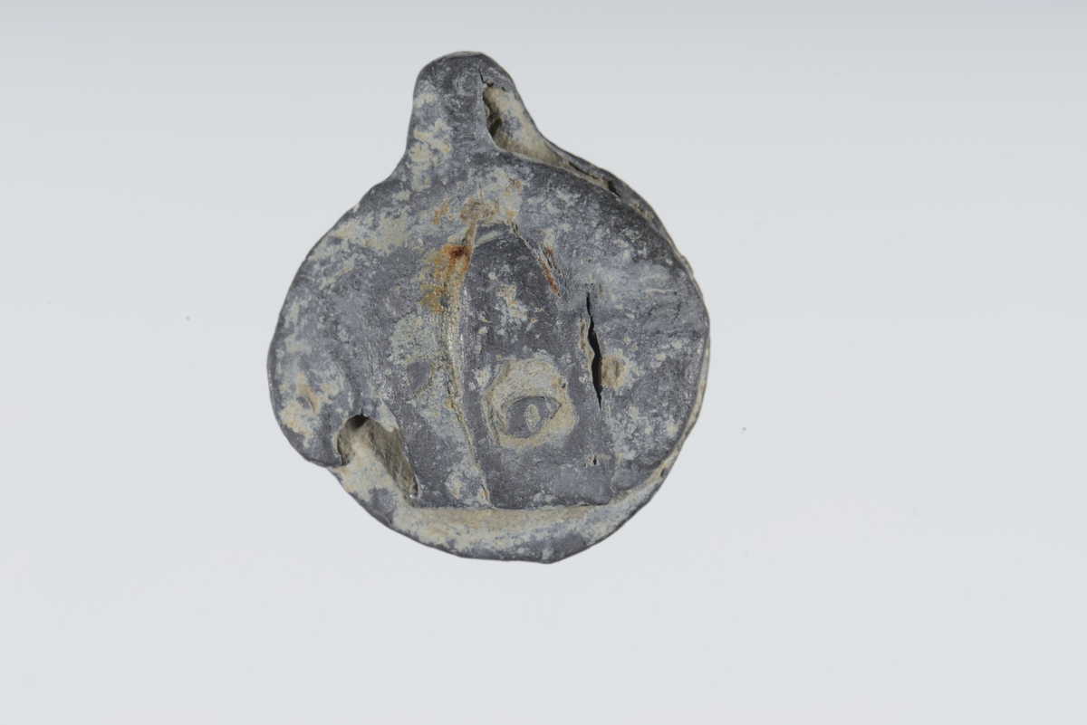 Vareplombe med begge plaketter, nagle og streng bevart. Gjenstanden er nedslitt men viser svake tegn til preg på begge plaketter. Preget er utydet. 