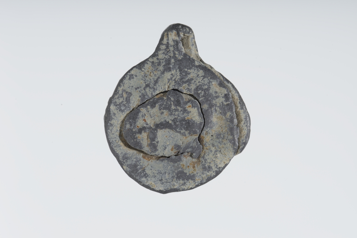 Vareplombe med begge plaketter, nagle og streng bevart. Gjenstanden er nedslitt men viser svake tegn til preg på begge plaketter. Preget er utydet. 