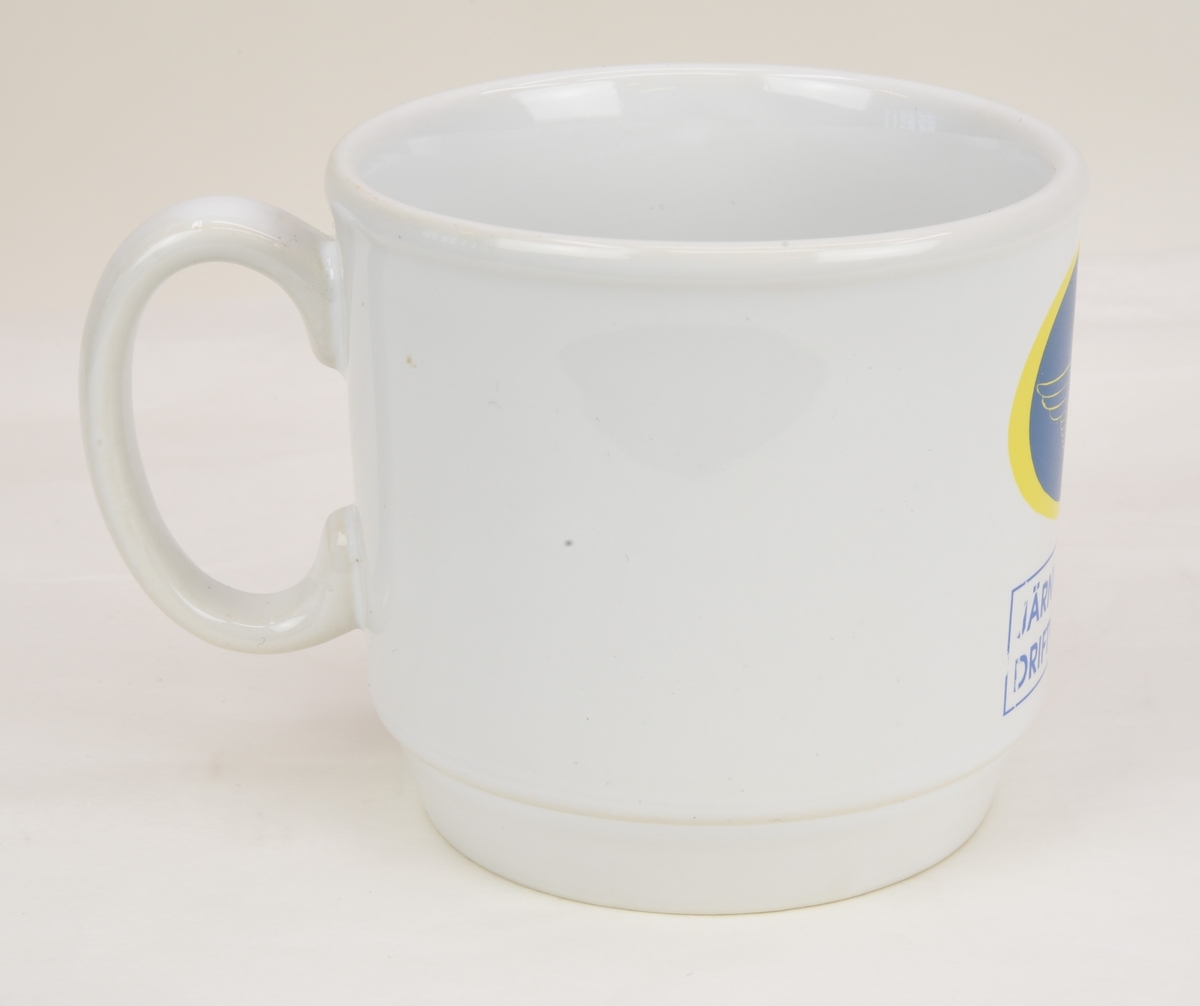 En vit kaffekopp av porslinmed tryckt motiv föreställande det bevingade samt krönta vinghjulet i en blå oval med gul kant. Nedanför texten "JÄRNVÄGARNAS DRIFTVÄRN" i en rektangel med en blå linje runtom.
