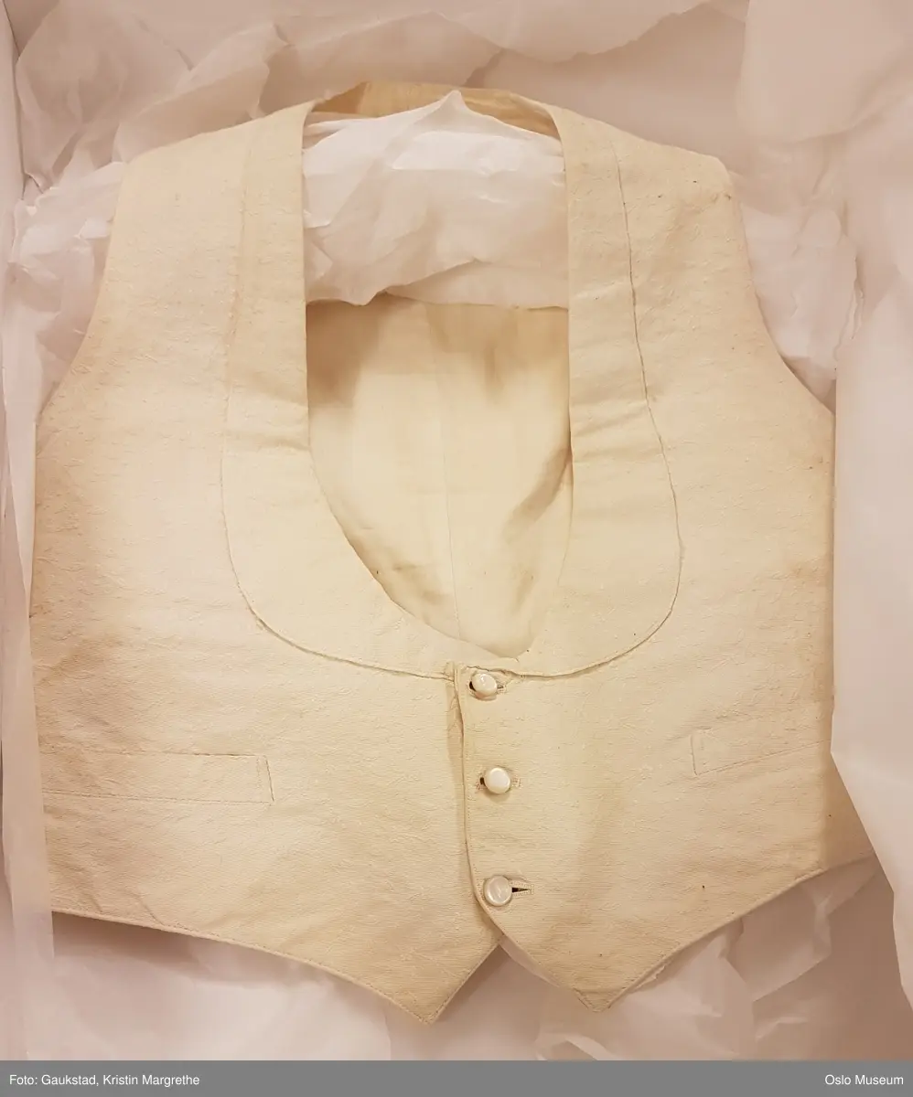 1: Vest i hvitt stoff med innvevde striper i brystparti. Knepping i form av 1 hvit knapp i perlemor, 3 mangler. Spenne bak.
2: Vest i hvitt stoff med innvevde ranker/blomster. Knepping foran med 3 hvite runde perlemor (?) - spenne bak