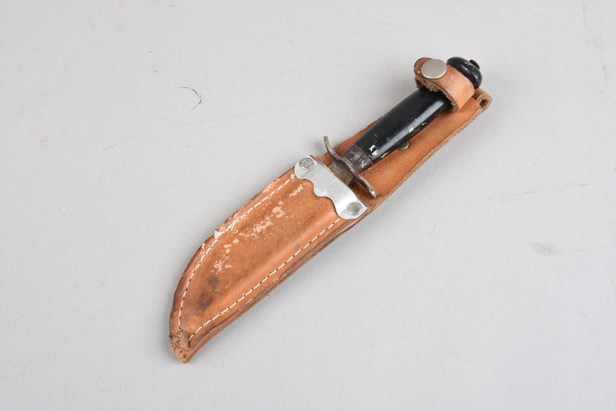 Speidarkniv med slire. Kniven har svart plasthandtak med tosidig fingerstopp mot knivblad, Skaftet har ovalt tverrsnitt. Slira er i lær, der delen som skjuler knivbladet er sydd saman med utvendig saum. Ved innstikket for knivbladet eit metallbeslag. Øvst på slira ei lerreim med trykknapp for feste rundt skaft. På baksida påsett ei lerhempe for beltefeste. Slira har prega dekor på framsida.