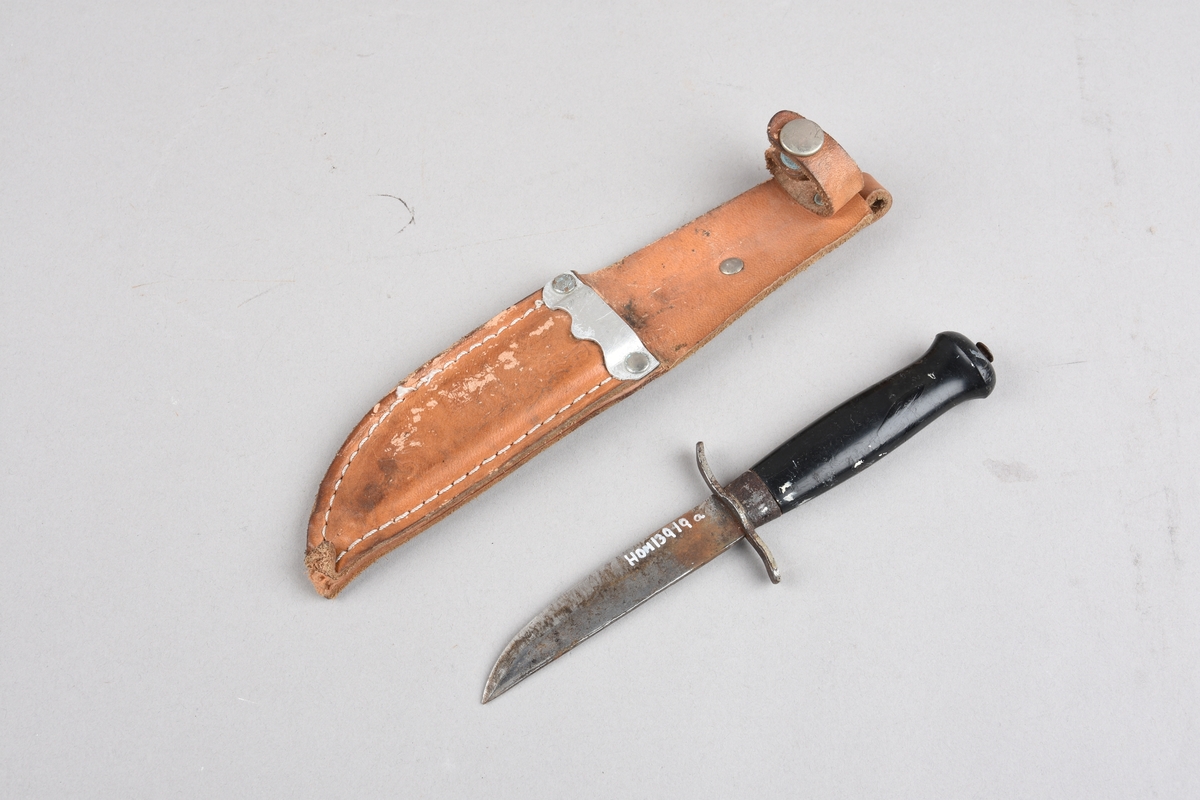 Speidarkniv med slire. Kniven har svart plasthandtak med tosidig fingerstopp mot knivblad, Skaftet har ovalt tverrsnitt. Slira er i lær, der delen som skjuler knivbladet er sydd saman med utvendig saum. Ved innstikket for knivbladet eit metallbeslag. Øvst på slira ei lerreim med trykknapp for feste rundt skaft. På baksida påsett ei lerhempe for beltefeste. Slira har prega dekor på framsida.