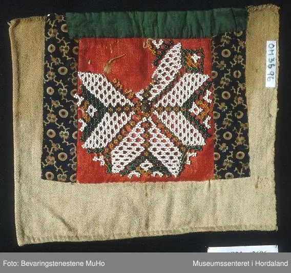 Rektangulær bringeduk med perlenett med åttebladrosemotiv sydd på raudt ullstoff. Perleveven er noko rakna og manglar eit blad. Midt i åttebladrosa er det sydd på ein knapp. Ullstoffet har fleire hol. Grøn silkeband oppe. Blått mønstrete bomullstoff på kvar side sydd på eit kypertvevd naturfarga stoff. Skittent på baksida. Det grøne bandet er sydd oppå eit anna rosafarga silkeband.