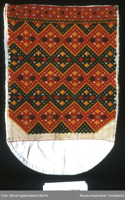 Brodert brystduk i repeterande mønster med sikksakk-border. Broderiet er rektangulært, bakstykket endar i ein halvrunding nedom broderiet. Stripete stoff på baksida.