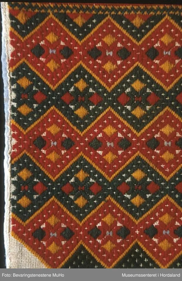 Brodert brystduk i repeterande mønster med sikksakk-border. Broderiet er rektangulært, bakstykket endar i ein halvrunding nedom broderiet. Stripete stoff på baksida.