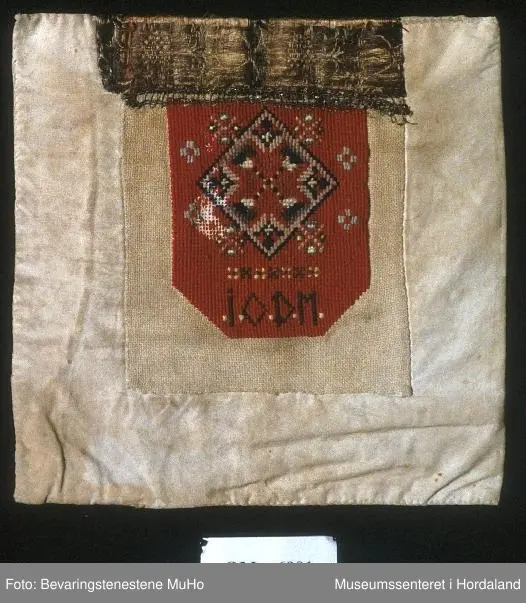 Rektangulær bringeduk sydd på kvadratisk bomullstoff. Broderiet er i halve og heile kross-sting sydd på linstoff. Raud hovudfarge. Initialane I O D M er sydd nederst på broderiet. Silkeband øvst med ei smal metallbord nedom. Silkebandet, metallborda og broderioet er veldig slitt.