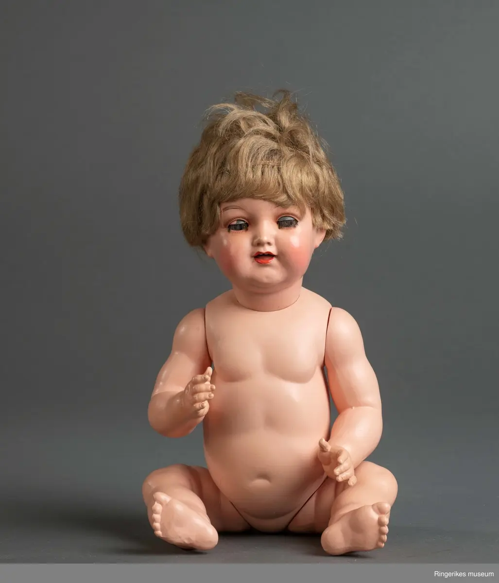 Dukke med babykropp, bøyde armer og ben, øyne som kan åpnes/lukkes og tykt hår. I kroppen lagt inn en boks som gir fra seg skrikelyd ved bevegelse.