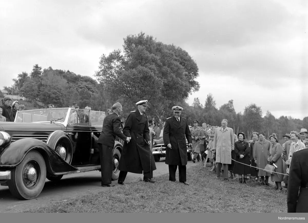 Bilen er en Buick 1935. (Info: Ivar Stav)    -Foto av Kong Haakon VII og Kronprins Olav i det de ankommer 1000-årsjubileet for slaget på Rastarkalv på Frei i Kristiansund kommune. Foto fra 1000-årsjubileet for slaget på Rastarkalv på Frei i Kristiansund kommune. Slaget på Rastarkalv var et slag i 955 mellom Eirikssønnene og Håkon Adalsteinsfostre. Eirikssønnene ledet en hær som bestod av norske og danske vikinger, tildelt av den første danske kongen, Harald Blåtann. Håkon Adalsteinsfostre vant slaget og tvang danskene til retrett. (Kilde: Wikipedia). Bildet er datert 1955. Fotograf er Nils Williams. Fra Nordmøre museums fotosamlinger.