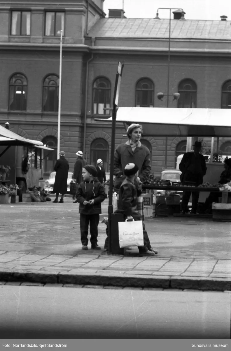 En kvinna och hennes söner väntar på bussen vid hållplatsen på Storgatan invid Stora torget. En av pojkarna håller en kasse från Centralaffären.