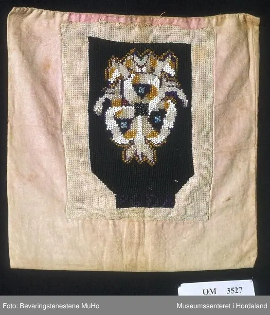 Form: rektangulær, ikkje kant oppe, men det er merke etter kant. Broderi sydd på stramei. Svart botn, påsydde perler i liljemotiv. Rosa, dobbelt bakstykke. Nedom det svarte broderiet er det ein svart brodert kant med initialer i lilla.
