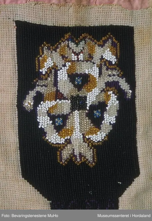 Form: rektangulær, ikkje kant oppe, men det er merke etter kant. Broderi sydd på stramei. Svart botn, påsydde perler i liljemotiv. Rosa, dobbelt bakstykke. Nedom det svarte broderiet er det ein svart brodert kant med initialer i lilla.