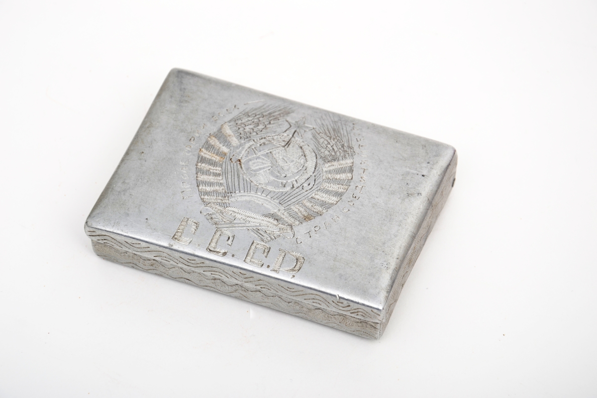Sigarettetui laget av sovjetisk krigsfange. Etui tilvirket av aluminium med pregstempeldekor. På lokket er det risset inn ulike symboler, blant annet hammer, sigd og den røde stjerne, sammen med kornaks  og tekst. På eskens underside er arbeideren (med fane) avbildet sammen med tekst. Det har vært dekor både på lokkets innside og i bunne av etuit, men disse er blitt risset over.