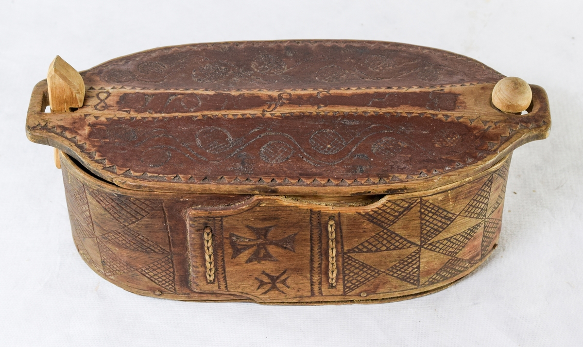 Furu, Furuspon bøyd kring oval botn og sydd manneh
med tæger. Skåret dekor, ranker og ruter. 2 jernkors mellom saumene i sida. Rep. fester for lokket. Rødmalt.